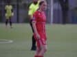 Female soccer player in a red uniform with number 9 stands on a green field; a referee in a neon yellow shirt is behind her.