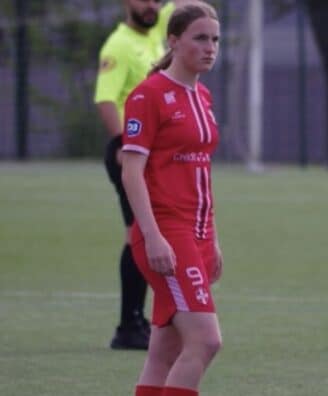 Female soccer player in a red uniform with number 9 stands on a green field; a referee in a neon yellow shirt is behind her.