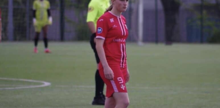 Female soccer player in a red uniform with number 9 stands on a green field; a referee in a neon yellow shirt is behind her.