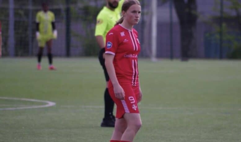 Female soccer player in a red uniform with number 9 stands on a green field; a referee in a neon yellow shirt is behind her.