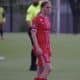 Female soccer player in a red uniform with number 9 stands on a green field; a referee in a neon yellow shirt is behind her.