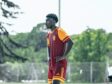 Male soccer player in maroon and orange kit stands with hands on hips on a grassy field during training.