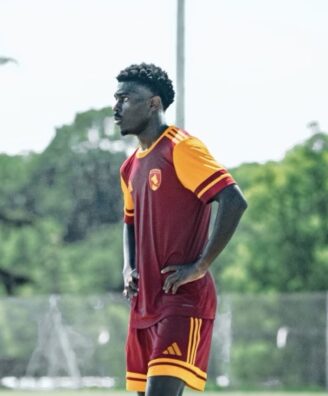 Male soccer player in maroon and orange kit stands with hands on hips on a grassy field during training.