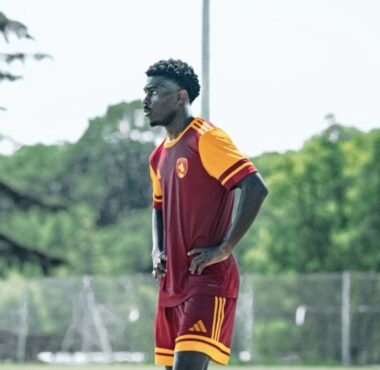 Male soccer player in maroon and orange kit stands with hands on hips on a grassy field during training.