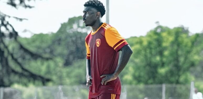 Male soccer player in maroon and orange kit stands with hands on hips on a grassy field during training.
