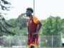 Male soccer player in maroon and orange kit stands with hands on hips on a grassy field during training.