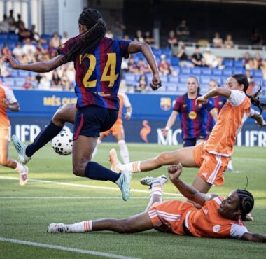 Soccer match scene: a player in a maroon-and-blue jersey (#24) leaps to kick the ball while players in orange defend nearby on a green field.