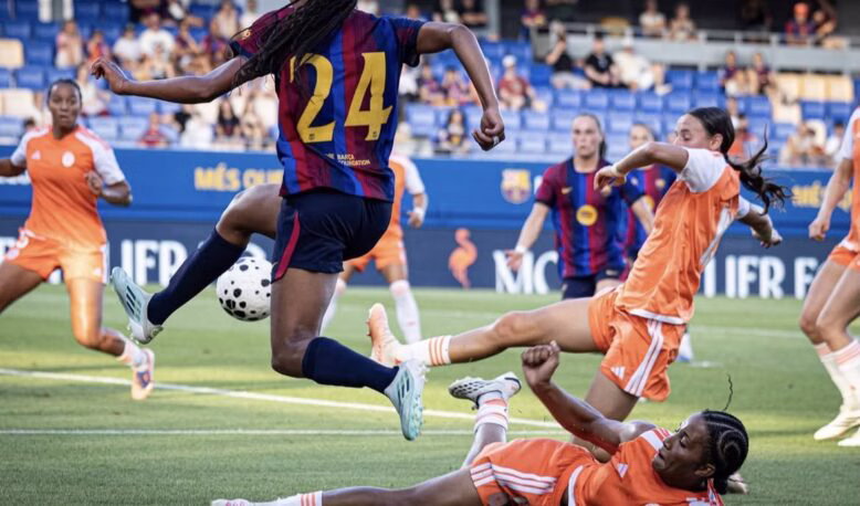 Soccer match scene: a player in a maroon-and-blue jersey (#24) leaps to kick the ball while players in orange defend nearby on a green field.