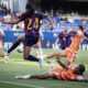 Soccer match scene: a player in a maroon-and-blue jersey (#24) leaps to kick the ball while players in orange defend nearby on a green field.