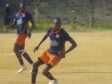 Lambert Sokoundjou | AllezPaillade.com Two soccer players in navy blue and orange uniforms compete on a dusty field; the foreground player wearing number 20 dribbles the ball as his teammate runs behind.