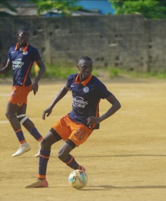 Lambert Sokoundjou | AllezPaillade.com Two soccer players in navy blue and orange uniforms compete on a dusty field; the foreground player wearing number 20 dribbles the ball as his teammate runs behind.