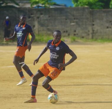 Two soccer players in navy blue and orange uniforms compete on a dusty field; the foreground player wearing number 20 dribbles the ball as his teammate runs behind.
