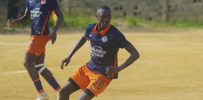 Lambert Sokoundjou | AllezPaillade.com Two soccer players in navy blue and orange uniforms compete on a dusty field; the foreground player wearing number 20 dribbles the ball as his teammate runs behind.