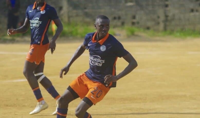 Two soccer players in navy blue and orange uniforms compete on a dusty field; the foreground player wearing number 20 dribbles the ball as his teammate runs behind.