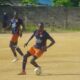 Two soccer players in navy blue and orange uniforms compete on a dusty field; the foreground player wearing number 20 dribbles the ball as his teammate runs behind.