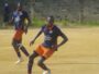 Two soccer players in navy blue and orange uniforms compete on a dusty field; the foreground player wearing number 20 dribbles the ball as his teammate runs behind.