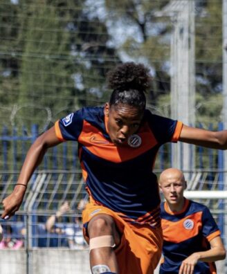 Screenshot | AllezPaillade.com Female soccer player in a navy and orange uniform mid‑stride, arms outstretched, on a grassy field during a match with a teammate in the background.