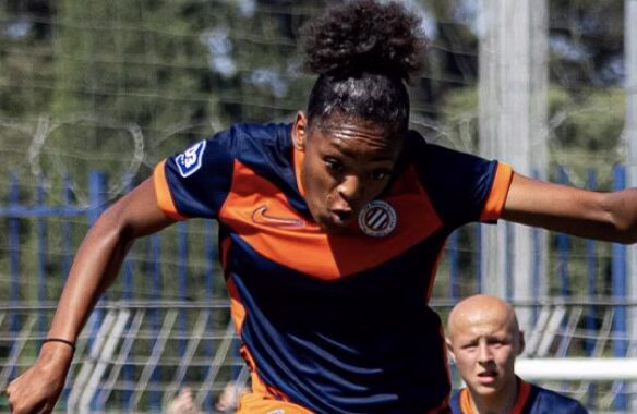 Screenshot | AllezPaillade.com Female soccer player in a navy and orange uniform mid‑stride, arms outstretched, on a grassy field during a match with a teammate in the background.