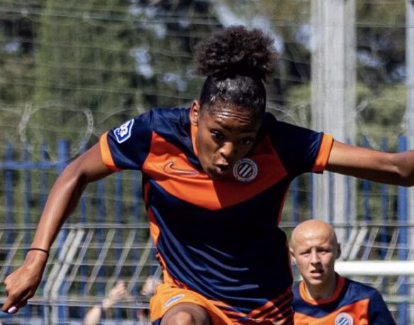 Screenshot | AllezPaillade.com Female soccer player in a navy and orange uniform mid‑stride, arms outstretched, on a grassy field during a match with a teammate in the background.