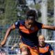 Female soccer player in a navy and orange uniform mid‑stride, arms outstretched, on a grassy field during a match with a teammate in the background.