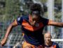 Female soccer player in a navy and orange uniform mid‑stride, arms outstretched, on a grassy field during a match with a teammate in the background.