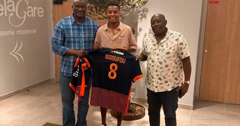 Three men pose indoors presenting a black and orange football jersey with name Issoufou and number 8.