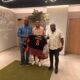 Three men pose indoors presenting a black and orange football jersey with name Issoufou and number 8.
