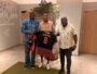 Three men pose indoors presenting a black and orange football jersey with name Issoufou and number 8.
