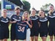 Six people in black sports uniforms standing on a grassy field, one woman in the center holds a blue jersey with the number 34 and text LES ARCEAUX, smiling at the camera.