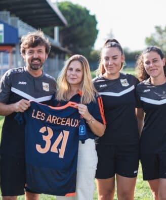 Six people in black sports uniforms standing on a grassy field, one woman in the center holds a blue jersey with the number 34 and text LES ARCEAUX, smiling at the camera.