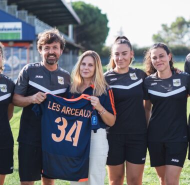 Six people in black sports uniforms standing on a grassy field, one woman in the center holds a blue jersey with the number 34 and text LES ARCEAUX, smiling at the camera.
