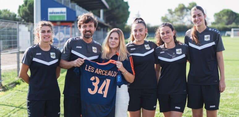 Six people in black sports uniforms standing on a grassy field, one woman in the center holds a blue jersey with the number 34 and text LES ARCEAUX, smiling at the camera.
