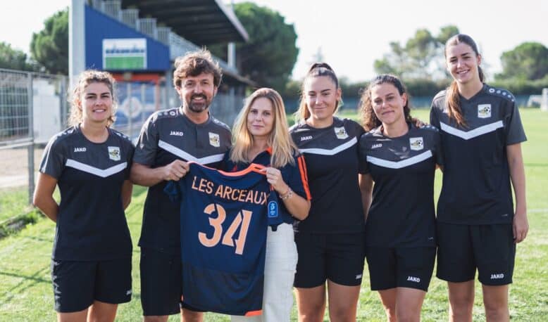 Six people in black sports uniforms standing on a grassy field, one woman in the center holds a blue jersey with the number 34 and text LES ARCEAUX, smiling at the camera.