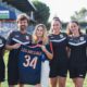 Six people in black sports uniforms standing on a grassy field, one woman in the center holds a blue jersey with the number 34 and text LES ARCEAUX, smiling at the camera.