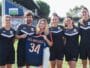 Six people in black sports uniforms standing on a grassy field, one woman in the center holds a blue jersey with the number 34 and text LES ARCEAUX, smiling at the camera.