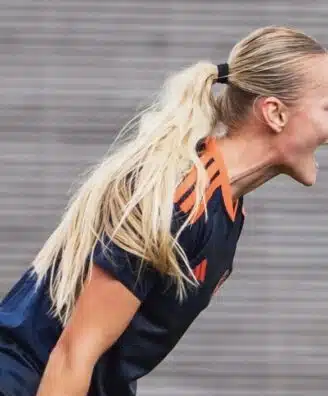 Female soccer player with a blonde ponytail, wearing a navy jersey with orange stripes, leaning forward and shouting.
