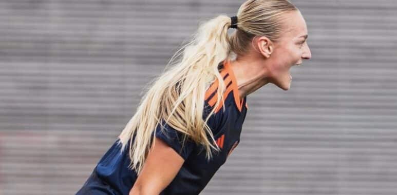 Screenshot | AllezPaillade.com Female soccer player with a blonde ponytail, wearing a navy jersey with orange stripes, leaning forward and shouting.