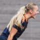 Female soccer player with a blonde ponytail, wearing a navy jersey with orange stripes, leaning forward and shouting.