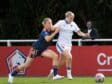 Two female soccer players compete for the ball on a sunny field, one in navy blue with orange trim and the other in white and blue, as they sprint side by side.
