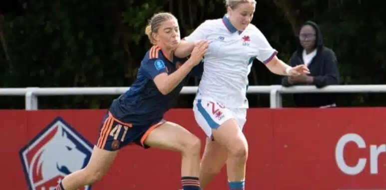 IMG_5892 | AllezPaillade.com Two female soccer players compete for the ball on a sunny field, one in navy blue with orange trim and the other in white and blue, as they sprint side by side.