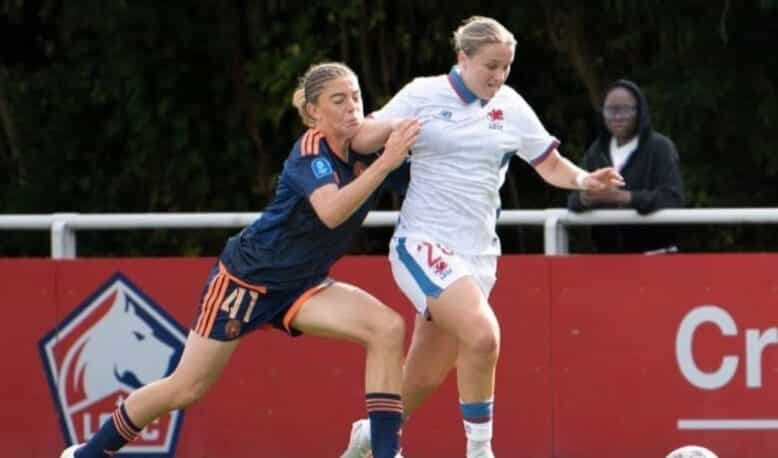 Two female soccer players compete for the ball on a sunny field, one in navy blue with orange trim and the other in white and blue, as they sprint side by side.