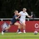 Two female soccer players compete for the ball on a sunny field, one in navy blue with orange trim and the other in white and blue, as they sprint side by side.