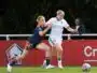 Two female soccer players compete for the ball on a sunny field, one in navy blue with orange trim and the other in white and blue, as they sprint side by side.