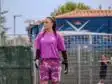 Female soccer goalkeeper in a pink jersey and patterned shorts stands on the field, wearing gloves and looking to the left with a fence and audience in the background.