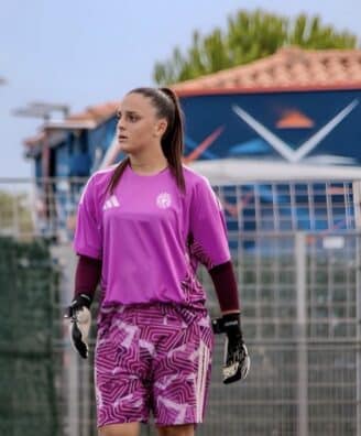 Female soccer goalkeeper in a pink jersey and patterned shorts stands on the field, wearing gloves and looking to the left with a fence and audience in the background.