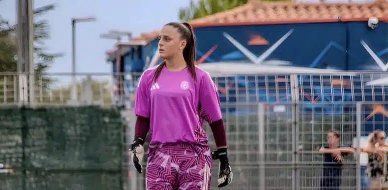Screenshot | AllezPaillade.com Female soccer goalkeeper in a pink jersey and patterned shorts stands on the field, wearing gloves and looking to the left with a fence and audience in the background.