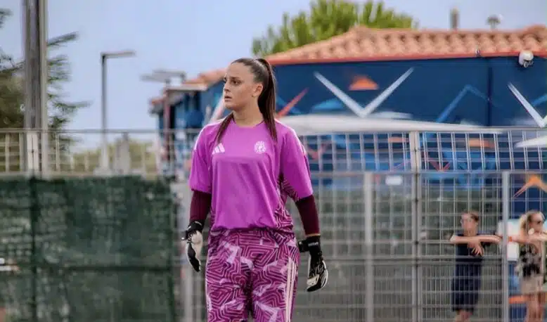 Female soccer goalkeeper in a pink jersey and patterned shorts stands on the field, wearing gloves and looking to the left with a fence and audience in the background.
