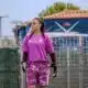 Female soccer goalkeeper in a pink jersey and patterned shorts stands on the field, wearing gloves and looking to the left with a fence and audience in the background.