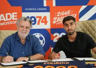 Two men sign documents at a table during a signing event, with a bright orange&ndash;blue graphic wall behind them.