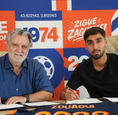 Two men sign documents at a table during a signing event, with a bright orange&ndash;blue graphic wall behind them.